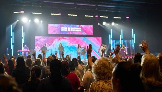 A view from behind a large crowd at a religious concert. Many people have their hands raised in worship towards a brightly lit stage where a band performs.